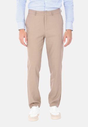 Uomo con pantaloni beige, camicia azzurra a righe e scarpe da ginnastica bianche, in piedi con le mani rilassate lungo i fianchi.