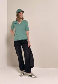 Frau mit grüner Mütze und Hemd, schwarzen Hosen und Turnschuhen, die eine schwarze Tasche hält, steht vor einer einfarbigen beigen Wand.