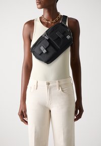 Borsa a tracolla nera in nylon con due tasche frontali, chiusura con zip e tracolla regolabile. Modello che indossa una canotta chiara e pantaloni color crema.