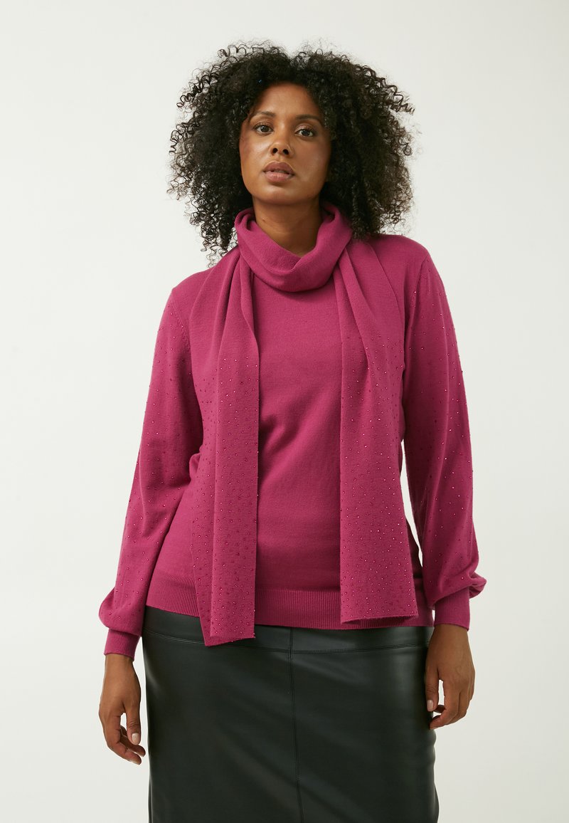 Fuchsia striktrøje med høj hals og lange ærmer. Læg stiloverslag med subtile perledetaljer. En sort lædernederdel fuldender outfittet.