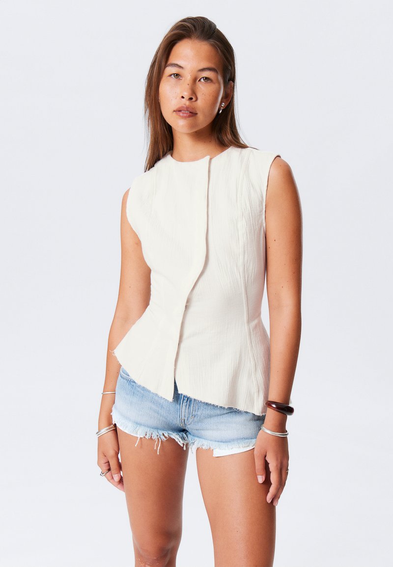 Top crema senza maniche con finitura tessurizzata, vita a peplo e orlo asimmetrico. Abbinato a pantaloni shorts in denim chiaro e strappato.
