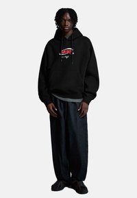 Čierna mikina s kapucňou a prednou kapsou, s grafickým logom v červenej a bielej farbe. Skombinovaná s voľnými tmavomodrými džínsy a čiernymi teniskami.