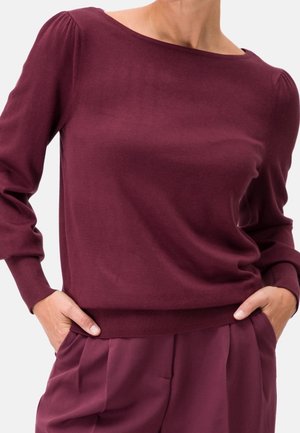 Femme portant un pull violet foncé à manches longues avec des épaules bouffantes et un pantalon assorti, les mains reposant négligemment dans les poches.