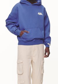 Sudadera de algodón azul marino con un bolsillo frontal y un logo blanco en el pecho, combinada con pantalones de cargo beige que tienen grandes bolsillos laterales.