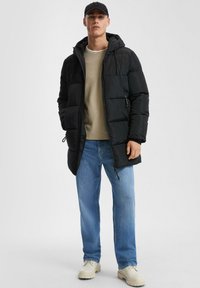 Mann in schwarzer Pufferjacke, beigem Pullover, blauen Jeans, kremfarbenen Stiefeln und schwarzer Mütze, der vor einem schlichten hellgrauen Hintergrund steht.