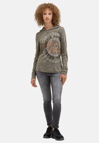 Olivegrüner Kapuzenpullover mit strukturiertem Design und bedrucktem Text in Schwarz und Orange. Kombiniert mit grauen Skinny-Jeans und schwarzen Sneakern.