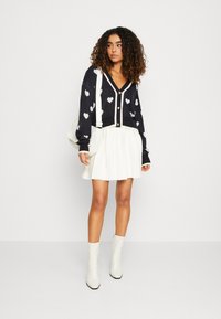 Navy vest met witte hartpatronen, gouden knopen, gecombineerd met een geplooide witte rok en kniehogere witte laarzen. Eenvoudige schoudertas.