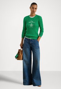 Camisola verde de malha com desenho de trança e logótipo, combinada com jeans azuis de perna larga, segurando uma mala castanha e um pequeno acessório verde.