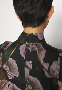 Blouse noire à motifs floraux avec un col haut, des manches bouffantes et un tissu transparent. Détails rayés remarquables et un dos en trou de serrure avec des boutons.