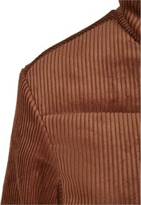 Bruin corduroy stof met verticale ribbels creëert een gestructureerde uitstraling. De afbeelding richt zich op het schoudergedeelte en de naaddetails.