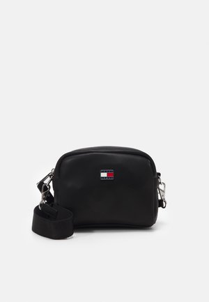 Bolso cruzado negro con una textura suave, forma redondeada, herrajes plateados y un pequeño logo con acentos en rojo, blanco y azul.