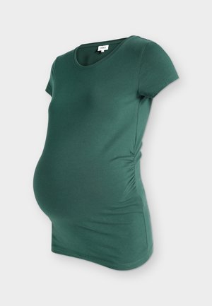 Έγκυος γυναίκα με πράσινο T-shirt, μαύρο φαρδύ παντελόνι, μαύρα σανδάλια, κρατώντας μια μεγάλη μαύρη τσάντα, στέκεται μπροστά σε απλό λευκό φόντο.