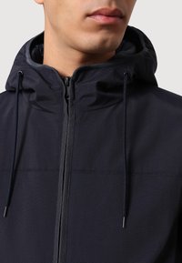 Giacca cappuccio blu scuro con zip frontale, dettagli con cordoncino e tessuto testurizzato. Fodera morbida visibile al colletto.