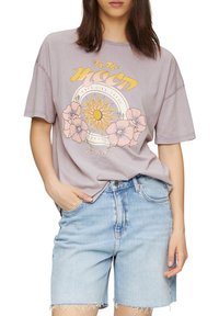 T-shirt oversize gris lavande avec imprimé graphique de soleil et de fleurs, associé à un short en denim bleu clair avec ourlet effiloché.