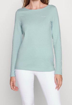 Kobieta w jasnoniebieskiej bluzce z długim rękawem z małym logo na piersi i białych dopasowanych spodniach na neutralnym tle.