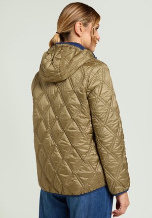 Kobieta o blond włosach spiętych w kucyk, ubrana w pikowaną, oliwkowozieloną kurtkę z kapturem i niebieskie dżinsy, widziana od tyłu i z profilu.