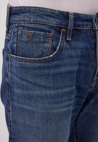 Primer plano de un bolsillo de jeans de mezclilla azul con la etiqueta Mavi Premium Denim y costuras naranjas visibles en una persona que lleva una camisa blanca.