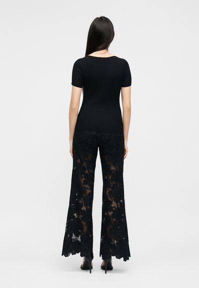 Γυναίκα με μακριά σκούρα μαλλιά που φοράει μαύρο ριμπ κοντομάνικο τοπ και μαύρο φαρδύ παντελόνι με φλοράλ δαντέλα, συνοδευόμενο από ψηλοτάκουνα, που κοιτάζει προς τα πίσω.