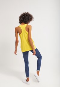 Gele tanktop met een racerback ontwerp, gecombineerd met getailleerde blauwe jeans en witte sneakers. De stof lijkt licht en textuur te hebben.