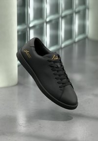 Μαύρο δερμάτινο αθλητικό παπούτσι με μαύρα κορδόνια και λογότυπο σε χρυσή καλλιγραφία στη πλευρά και στη γλώσσα, να αιωρείται πάνω από γκρι γυαλιστερό πάτωμα.