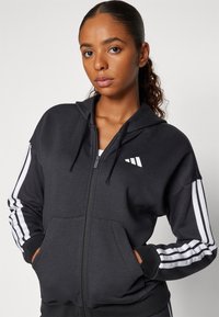 Black zip-up hoodie with a hood, features white stripes along sleeves, front pockets, and a small logo on the chest. Soft fabric texture.
