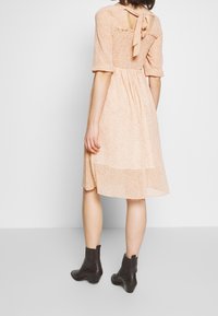 Žena v midi šatech světle broskvové barvy s vázáním na zádech a řaseným živůtkem, obutá do černých kotníkových bot, stojí proti jednobarevnému pozadí.