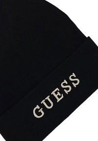 Beanie in maglia nera con risvolto e testo bianco ricamato "GUESS". Texture liscia, forma rotonda e senza motivi visibili.