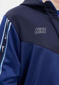 Tumši zils Nike kapuča ar rāvējslēdzēju, kuram ir gaišāka zila akcenti un izteikts balts logo uz krūtīm. Mīksta, teksturēta auduma.