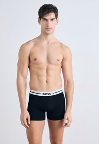 Čierne boxerky s bielym elastickým pásom, na ktorom je logo "BOSS". Vyrobené z bavlny, majú hladkú textúru a priliehavý strih.