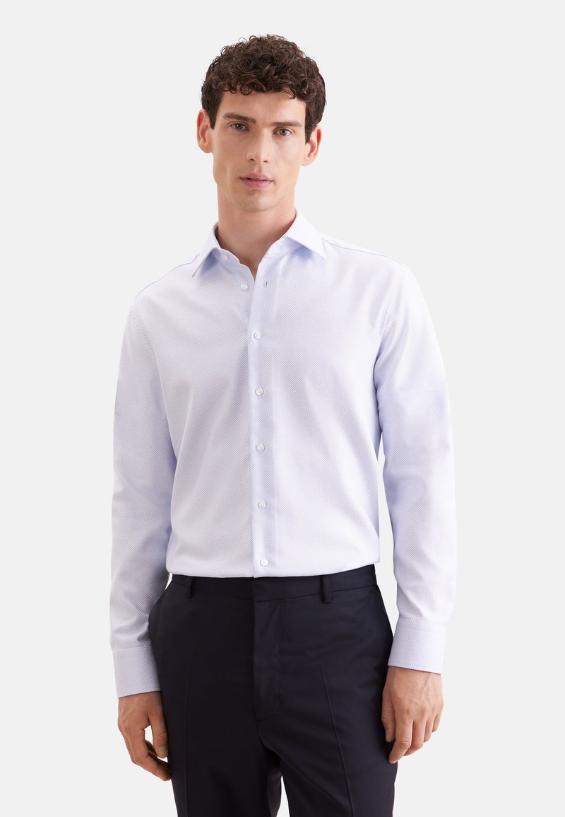 Homme aux cheveux courts et bouclés portant une chemise à manches longues boutonnée bleu clair à motif et un pantalon sombre, debout devant un fond uni.
