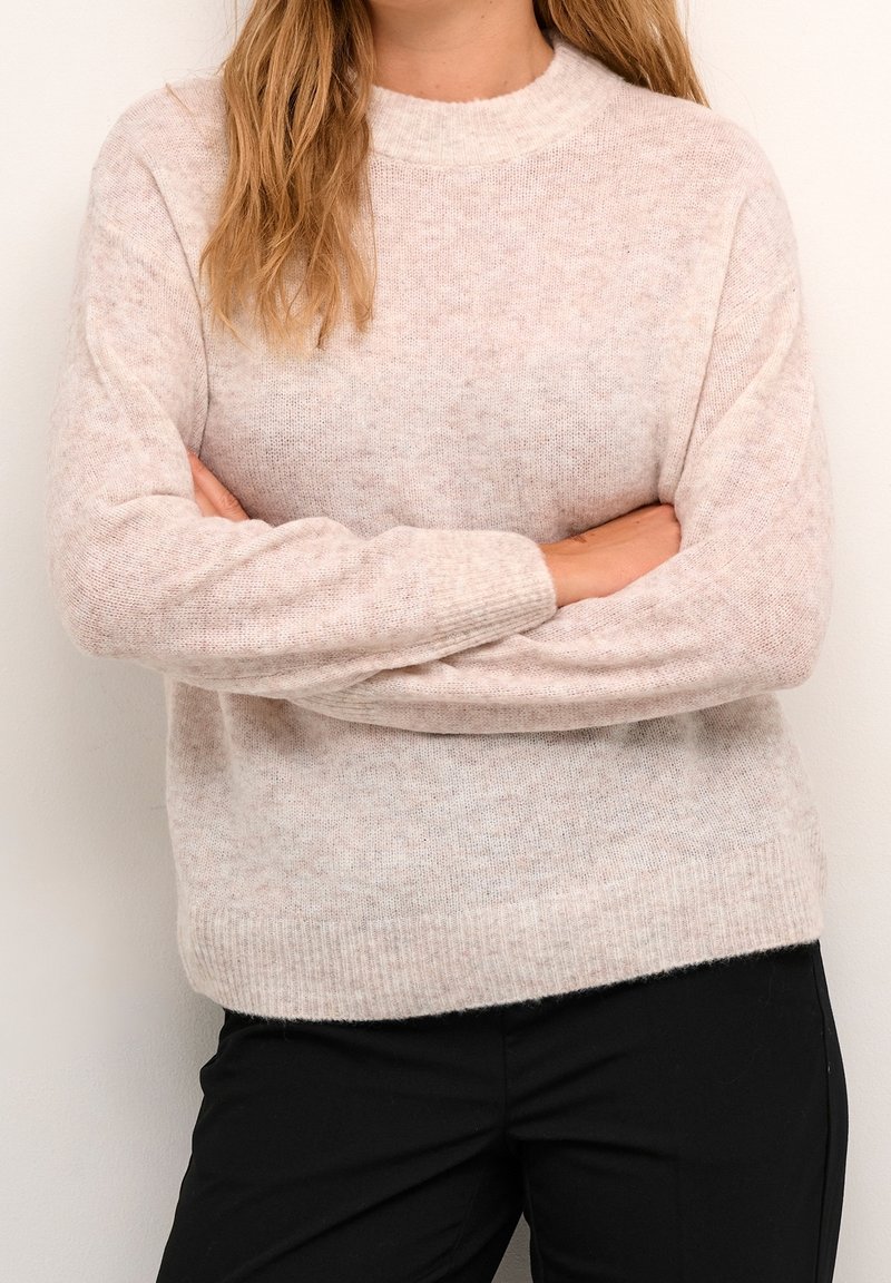 Person mit langen, hellbraunen Haaren, die einen hellrosa Strickpullover und schwarze Hosen trägt, steht mit verschränkten Armen vor einer weißen Wand.