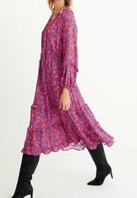 Robe longue à motif floral dans des nuances de violet et de rose, en tissu léger, avec des volants superposés, des manches longues avec des poignets élastiques, associée à des bottes à talons noires.