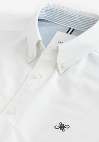 Weißes Hemd mit Button-Down-Kragen, ausgestattet mit einem hellblauen gestreiften Innenfutter und einem kleinen schwarzen gestickten Logo auf der Brust.