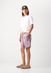 Shorts de bain violet clair avec des motifs de parasols multicolores et de vagues, taille élastique et poches latérales. Associé à un t-shirt blanc et une casquette brune.