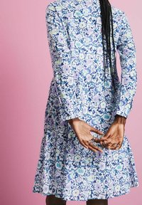 Personne avec de longues tresses portant une robe à manches longues à motifs floraux bleu, blanc et violet, se tenant les mains jointes derrière le dos sur un fond rose.