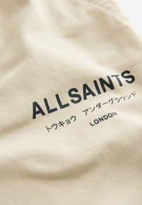Beige tyg med ett svart tryckt logo "ALLSAINTS" och text på japanska och engelska. Mjuk textur med synliga söm detaljer.