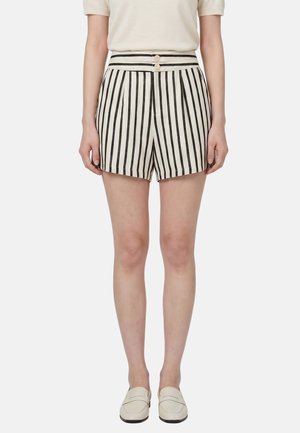 Donna che indossa pantaloncini a righe verticali bianchi e neri a vita alta con due bottoni, una maglia beige e mocassini crema, in piedi rivolta in avanti.