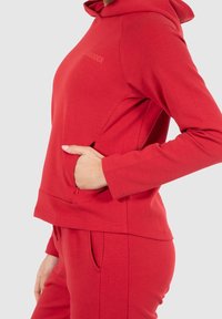 Sweatshirt à capuche rouge avec une finition texturée, doté d'une poche avant et de manches longues. Coupe décontractée, associé à un bas rouge assorti.