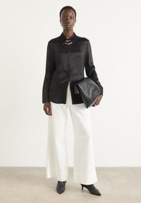 Camicia nera in satin a maniche lunghe, pantaloni bianchi a gamba larga, décolleté nere a punta, con una pochette in pelle nera, indossando una collana a catena.