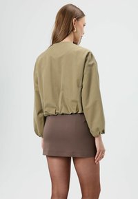 Olijfgroene blouse met lange mouwen en geplooide zoom, gecombineerd met een bruine, aansluitende mini-rok, tentoongesteld tegen een effen achtergrond.