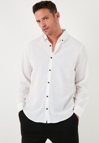 Camisa blanca de manga larga hecha de un tejido ligero, con cuello tipo camisa abotonado y botones negros en el frente, con una textura suave.