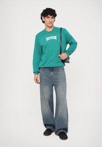 Teal sweatshirt med hvit "BARROW" tekst, kombinert med vide blå jeans og svarte sko. Modell med en liten svart veske.