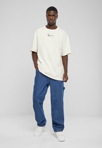 Krämmig t-shirt med kort ärm och svart logotyp, kombinerad med blå baggy denimjeans och vita sneakers. Enkel och avslappnad design.