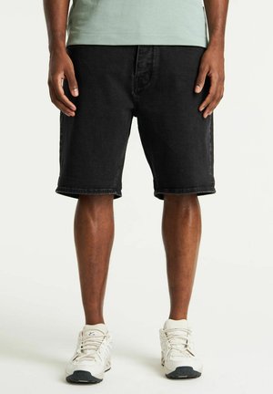 Pantalones cortos de mezclilla negros con un ajuste holgado, dobladillo enrollado y bolsillos delanteros, combinados con zapatos deportivos blancos y una camiseta verde.