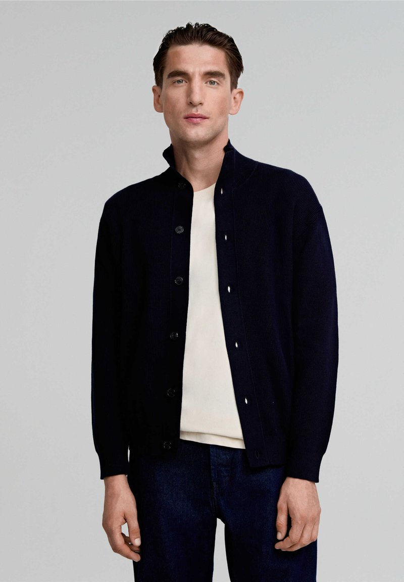 Cardigan di un blu navy scuro con texture a costine, chiusura con bottoni e colletto alto, indossato sopra una camicia chiara e jeans di denim scuro.