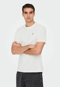White cotton t-shirt with short sleeves, round neckline, and small blue floral embroidery on the left chest. Worn with gray jeans.