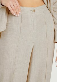 Beige linnen broeken met een hoge taille, voorzien van een sluiting met knopen aan de voorkant en subtiele plooien; gladde textuur met een lichtgewicht, ademend gevoel.