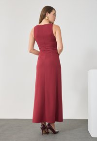 Rotes, ärmelloses Maxikleid mit tailliertem Oberteil, ausgestelltem Rock und einem unsichtbaren Reißverschluss am Rücken, kombiniert mit glänzenden bordeauxfarbenen High Heels.