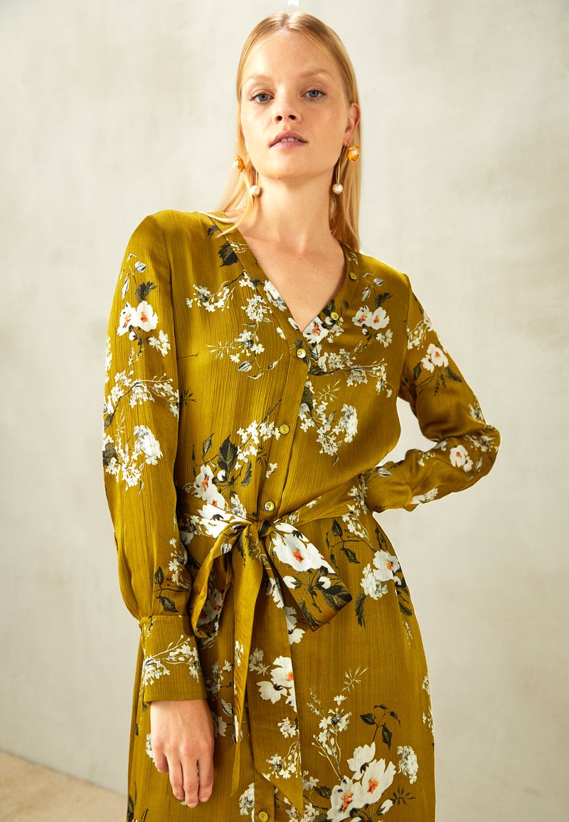 Vestido floral verde com mangas longas, decote em V e cintura ajustada com um cinto combinando. Apresenta detalhes em branco e escuros.