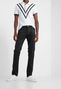 Bílá polokošile s černými chevronovými pruhy, černé džíny a bílé tenisky. Polokošile má límec a trojitý knoflíkový rozparek.